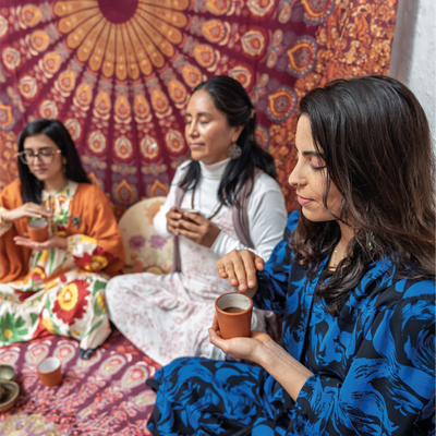 Three women sitting on a colorful rug, holding cups and engaged in conversation.